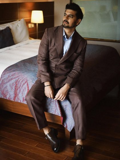 Tahir Raj Bhasin seated on a bed, dressed in a sharp suit. This portrait blends formal wear with a more casual, intimate setting.