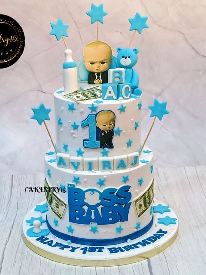 A classic two-tier Boss Baby cake for a first birthday. This design features a fondant Boss Baby topper, a teddy bear, ABC blocks, and edible dollar bills.