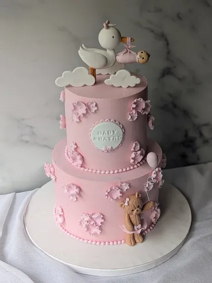 A classic pink baby shower cake. This two-tier design features a stork delivering a baby, a teddy bear, and delicate floral details.