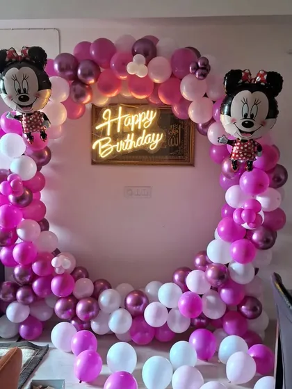 A Minnie Mouse themed birthday party decoration. I used a circular balloon arch with pink, purple, and white balloons, Minnie Mouse foil balloons, and a central 'Happy Birthday' neon sign.