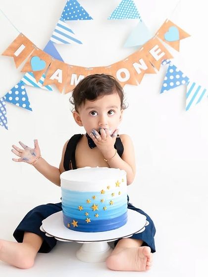 A classic cake smash pose with a blue ombre cake, set against a festive "I am one" banner.