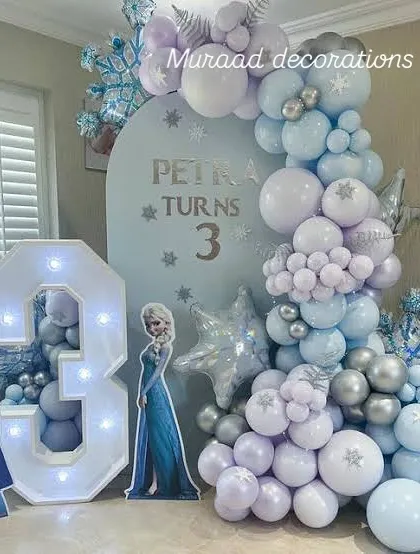 A winter wonderland for a third birthday with this Frozen theme. The decoration features a light-up marquee number '3', cutouts of Anna and Elsa, and a balloon garland with snowflake details.