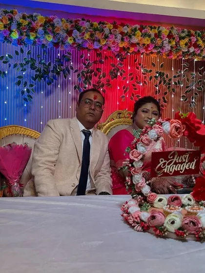 A happy couple during their engagement ceremony on a stage I decorated. The backdrop features a multi-colored floral border and fairy lights.