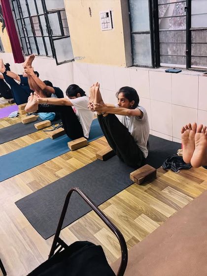 Students practice a variation of Navasana (Boat Pose), building core strength and balance. The focus and effort are clear on their faces.