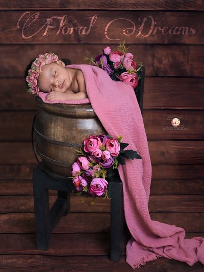 A sleeping beauty surrounded by flowers. This newborn is posed in a rustic wooden pot, draped in a soft pink blanket with floral accents, creating a beautiful, artistic portrait.
