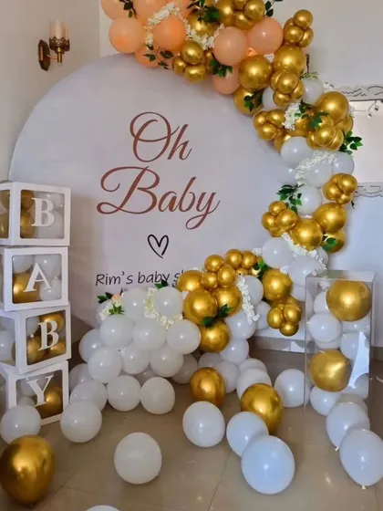 An elegant "Oh Baby" shower theme in white and gold. This decoration features a partial balloon arch over a custom backdrop, with transparent 'BABY' blocks filled with matching white and gold balloons.