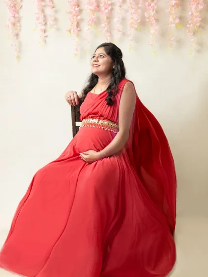 A radiant, high-key maternity portrait. The bright background, flowing red gown, and floral elements above create an atmosphere of pure joy and celebration for the mom-to-be.