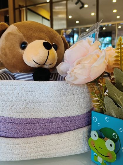 A cute teddy bear in a woven rope basket, part of a larger hamper curation for a child.