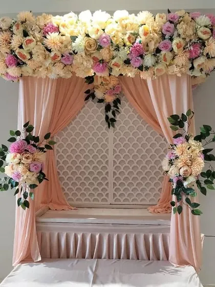 We create elegant and sophisticated decorations like this one, using a soft pastel color palette. The arrangement features a dense arch of artificial peach and cream flowers over matching drapes and a white lattice panel, offering a clean and modern aesthetic.