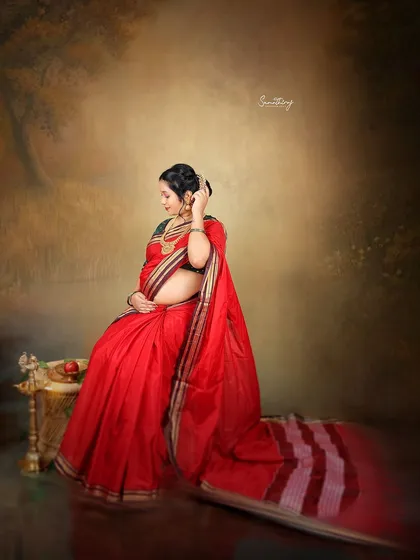 The vibrant red of this saree symbolizes love and new beginnings. This portrait, set against a painterly backdrop, captures a moment of quiet reflection and the timeless grace of an expecting mother.