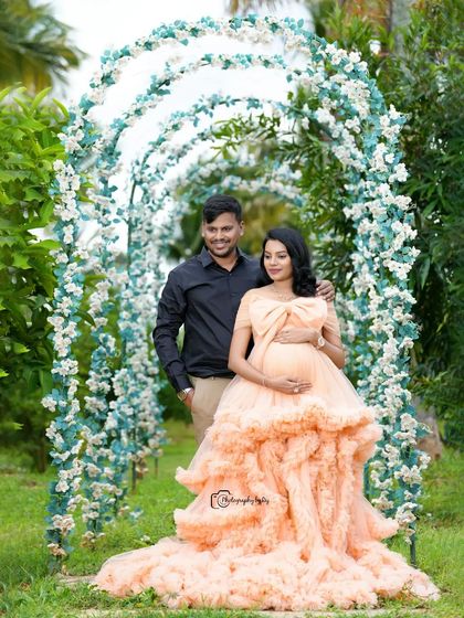 A stunning couple's portrait under a floral archway. The expecting mother looks radiant in a ruffled peach gown, creating a picture-perfect moment.