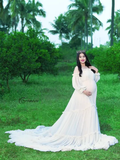 A portrait of pure elegance. The mother-to-be looks regal in a flowing white gown and a delicate crown, set against the natural green backdrop of our exclusive outdoor location.
