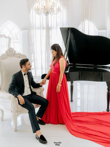 Elegance and romance personified. The groom seated in a classic armchair, holding his bride's hand as her stunning red gown trails beside them, set against a grand piano.