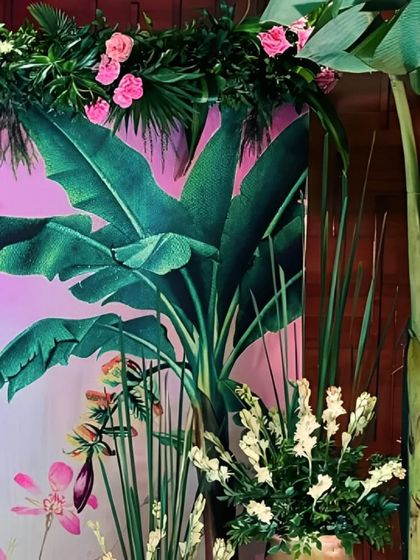 A sophisticated decor detail, combining a printed backdrop of tropical leaves with real banana tree stems and arrangements of white and pink flowers.