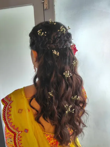This half-up, half-down hairstyle for a Haldi ceremony is both beautiful and practical. It features a braided crown and soft waves, decorated with baby's breath and a single bright flower for a pop of color.