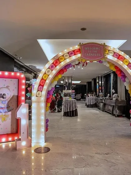 A grand carnival entrance archway for an indoor event. The arch is decorated with colorful balloons and marquee lights, flanked by light-up signs and a friendly elephant prop to welcome guests to the celebration.