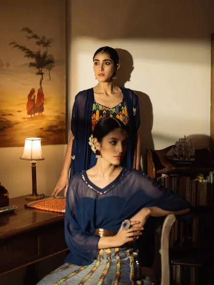 A moody, atmospheric shot from a lookbook, featuring two models in a dimly lit, studious room. The lighting is cinematic, highlighting the deep blue tones of the outfits.