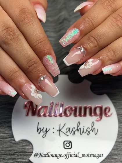 An elegant coffin-shaped nail set. The design includes a sheer pink base, white V-tips, iridescent glitter, and 3D flowers with rhinestone centers.