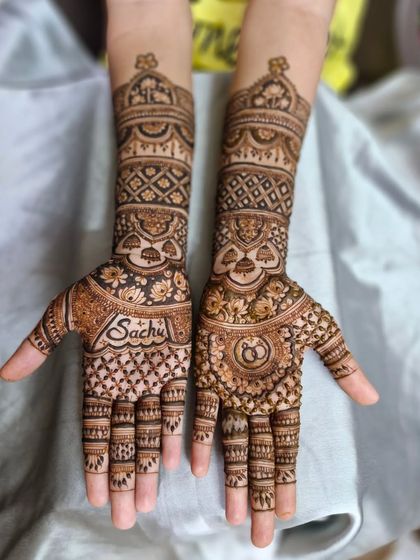 A personalized engagement mehendi with the name 'Sachu' written on the palm. The other hand features the classic symbol of interlocking rings.