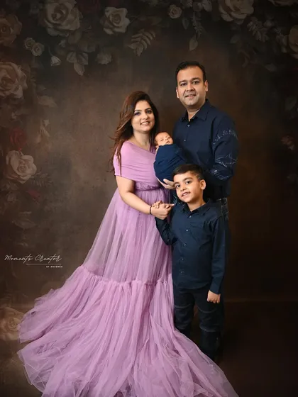 A beautiful family of four with their newborn. The mother's flowing lavender gown adds a soft touch of color to this elegant portrait against a dark, floral-painted backdrop.