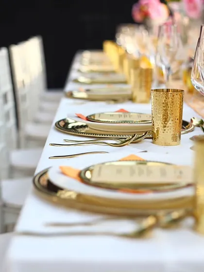 A long dining table set with gleaming gold-plated cutlery and glasses against crisp white linens. This modern and luxurious setup is perfect for an elegant wedding reception or corporate gala.
