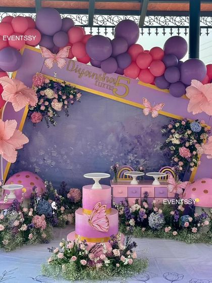 A whimsical floral and butterfly theme for a 5th birthday, using a stunning color palette of purple and pink. The setup features a custom backdrop, oversized paper flowers, and magical mushroom props.
