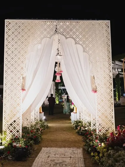 An ethereal entrance for a timeless wedding. A grand arch with intricate jaali work and soft white drapes, flanked by lush pink and white florals, welcomes guests into an enchanting world.