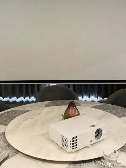 A clean and minimalist projector setup. This Optoma projector is placed on a table, projecting onto a large screen, showing how easy it can be to create a home cinema in any room without complex mounting.