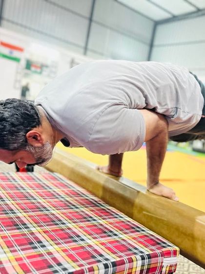 An adult student practices a peacock pose (mayurasana) on the balance beam. This advanced arm balance builds incredible wrist, forearm, and core strength.