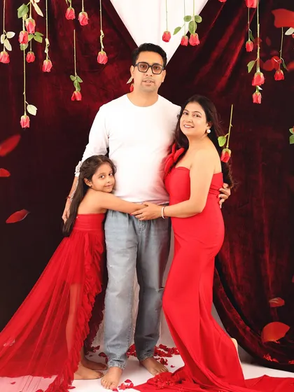 A classic family pose against a romantic red backdrop with hanging roses. The daughter hugs her father while the expecting mother looks on, creating a warm and connected family portrait.