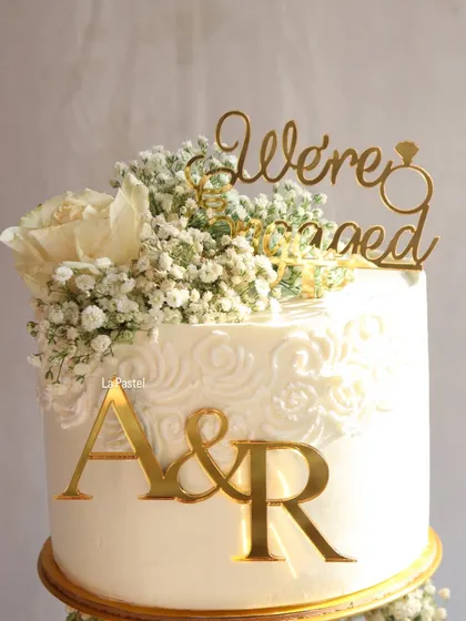 A close-up of the top tier, showing the custom 'We're Engaged' topper, personalized initials, and delicate lace-like piping.