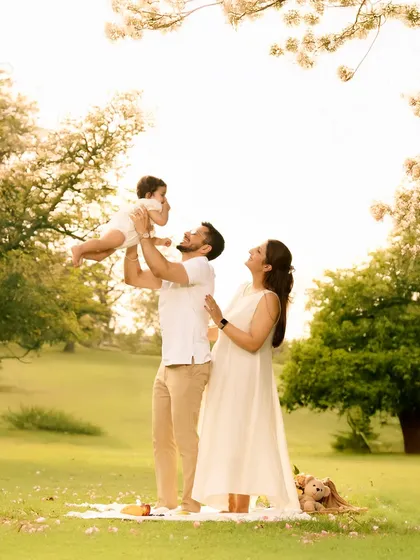 A beautiful, sunlit moment of a family enjoying a picnic in a lush green park. The father joyfully lifts his baby into the air as the mother looks on with a smile, perfectly capturing the warmth and happiness of candid family life.