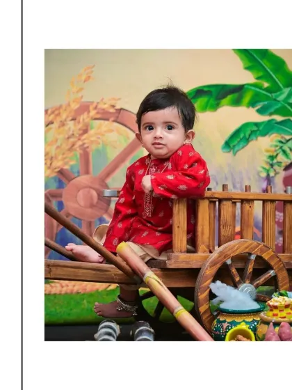A handsome little boy in a red kurta sits on a decorative bullock cart for a Sankranthi themed shoot. The backdrop of banana leaves and harvest elements creates a perfect festive atmosphere.