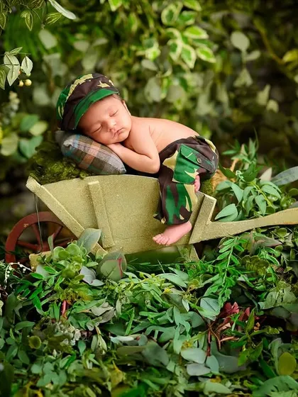 A baby dressed in camouflage rests in a wheelbarrow overflowing with lush greenery, creating a vibrant and adventurous jungle explorer scene.