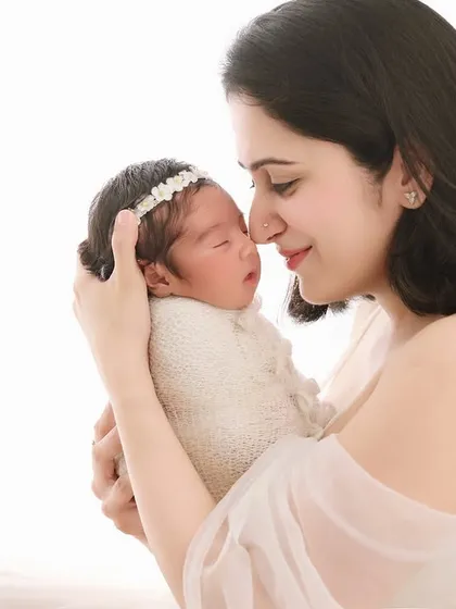 A beautiful mother-and-child portrait against a clean, bright background. The focus is entirely on the loving gaze between the mother and her sleeping newborn, wrapped in a textured knit outfit.