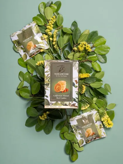 A creative flat lay of Taste & Beyond cookie packaging arranged on a bed of fresh green leaves. This styling emphasizes the natural ingredients and freshness of the product.