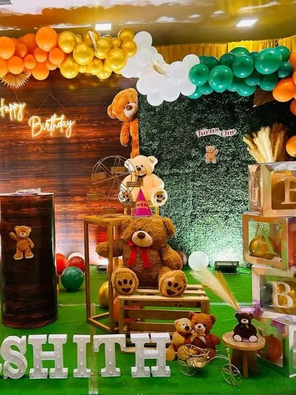 A charming teddy bear-themed birthday party. The setup uses a wood-paneled backdrop, a greenery wall, and an orange, green, and gold balloon arch to create a warm, rustic feel.