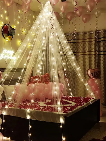 To create an unforgettable romantic birthday surprise, I design complete room decorations. This setup features a canopy draped with fairy lights over the bed, a floor and bed covered in rose petals, and pink balloons to create a warm and intimate atmosphere.
