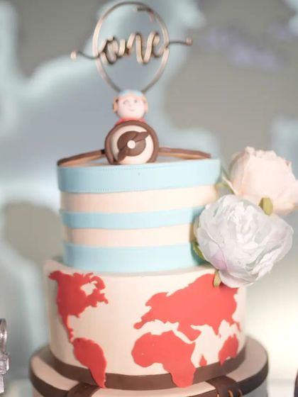 A close-up of a charming travel-themed cake. The design includes a world map, an airplane topper, and a "one" sign, making it a perfect centerpiece for a first birthday adventure.