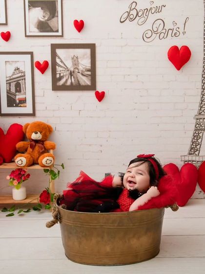 A happy baby girl laughs with delight in a tub, surrounded by red and black balloons in a "Bonjour Paris" Valentine's theme.