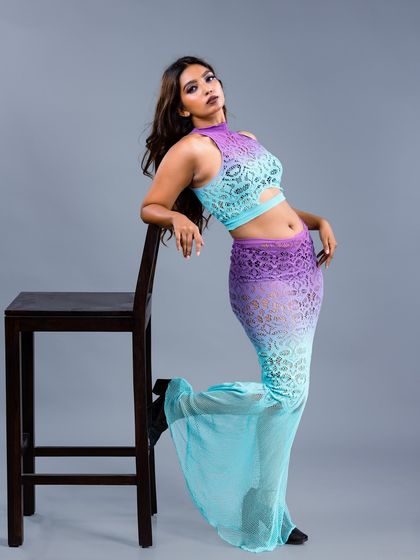 A model in a unique purple and blue ombre lace two-piece outfit, leaning against a stool. The colors and texture make this a standout look for an editorial shoot.