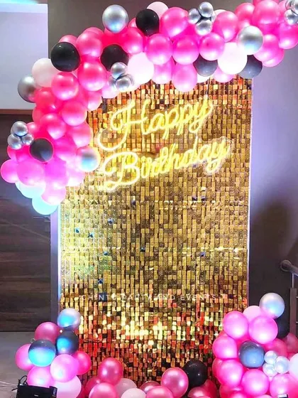 A modern and glamorous birthday photo booth with a gold shimmer sequin wall. The setup is completed with a custom neon sign and a vibrant pink and black balloon arch, perfect for chic celebrations.