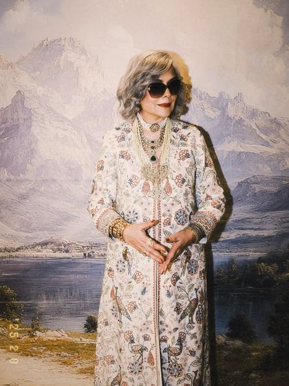 The full look for Zeenat Aman against a scenic backdrop. The long, printed jacket paired with wide leg trousers and layers of traditional jewelry creates an aura of effortless royalty.