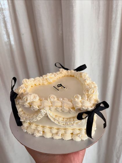 A beautiful vintage-style cake in a unique rectangular heart shape. The off-white buttercream is detailed with intricate piping and finished with small black bows for a classic look.