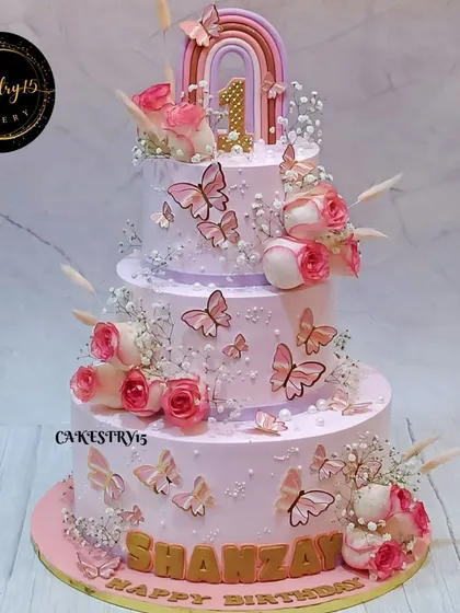 A grand three-tier butterfly-themed cake for a first birthday. This stunning creation is adorned with fluttering butterflies, real pink roses, and a fondant rainbow topper.