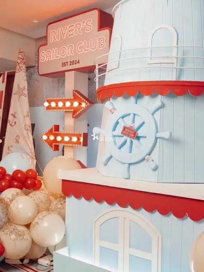 A custom-built lighthouse structure for 'River's Sailor Club,' a nautical-themed first birthday. This prop, complete with a ship's wheel and marquee signage, served as a stunning focal point for the celebration.