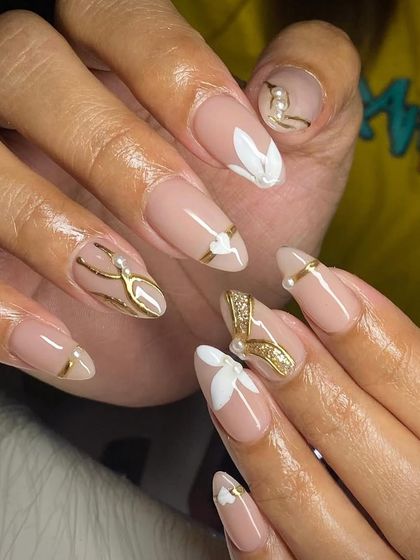 Elegant and sophisticated. These almond nails feature a nude base with delicate white floral art, gold lines, and pearl accents.