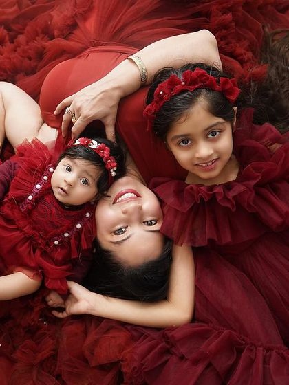 A creative and loving portrait of a mother with her two daughters. Lying on a bed of red fabric, she is surrounded by her girls, creating a beautiful image of sisterly and motherly love.