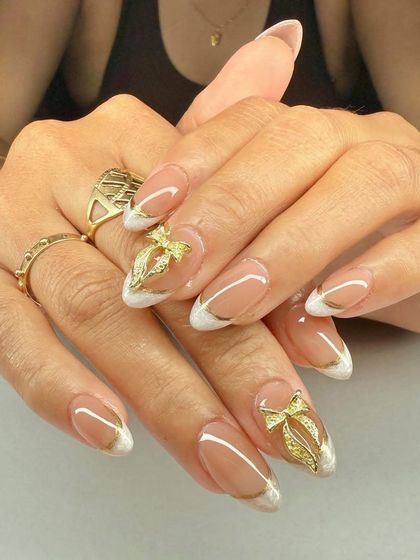 A glamorous French manicure with a twist. These almond nails feature gold chrome tips and are adorned with large, sparkling 3D gold bow charms.