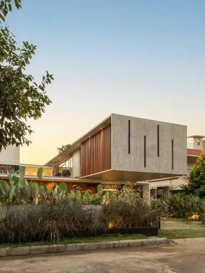 The Long House viewed from the garden at dusk. The cantilevered concrete volume appears to float above the timber-clad ground floor, with the surrounding landscape and wild grasses softening its modern lines.